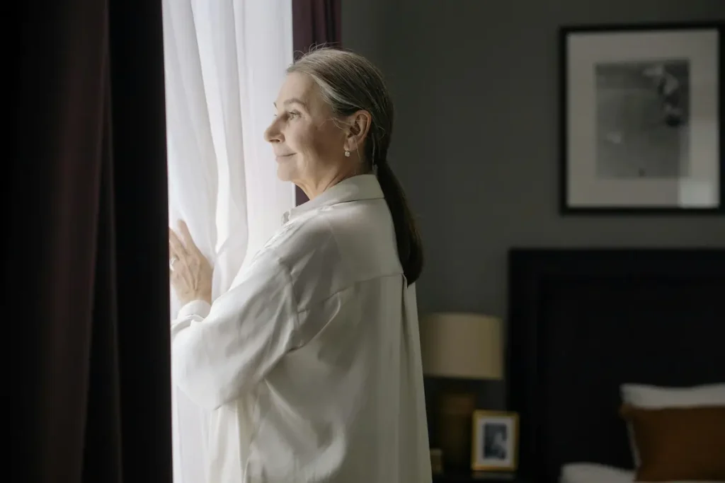 Mujer madura con expresión de tranquilidad mirando por la ventana con una sonrisa tranquila.